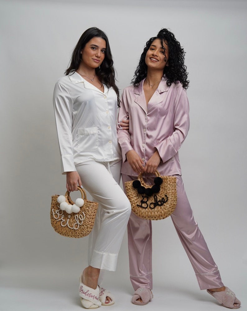May include: Two people wearing matching satin pajamas, one white and one pink, holding straw bags with the words "Bride" and "Babe" on them. They are both wearing matching slippers with the words "Bride" and "Bridesmaid" on them.