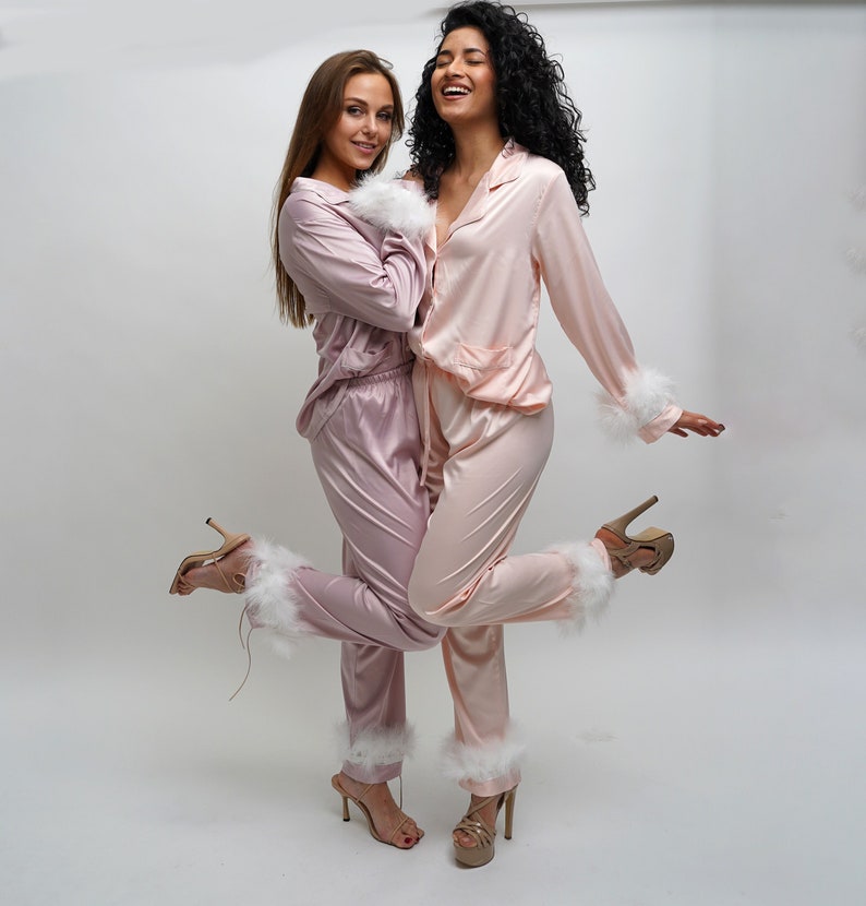 May include: Two women wearing matching pink satin pajamas with white feather trim. They are both smiling and looking at the camera. The pajamas have a relaxed fit and are perfect for lounging around the house.