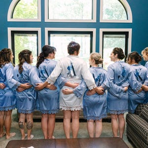 May include: Eight women in matching light blue satin robes with lace trim stand in a row, facing away from the camera. The robes have white embroidery on the back. The women are in a room with a large window behind them.