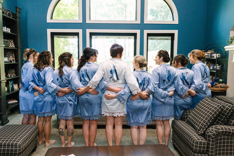 May include: Eight women in matching light blue satin robes with lace trim stand in a row, facing away from the camera. The robes have white embroidery on the back. The women are in a room with a large window behind them.