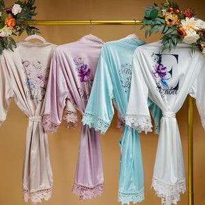 May include: Four satin robes with lace trim, hanging on a gold rack. The robes are in beige, lavender, light blue, and white. The robes have floral designs and text that reads "Maid of Honor", "Bride", "Mrs. & Mr." and "E Isabel".