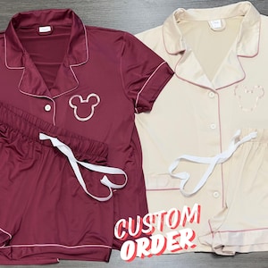 May include: Two sets of pajamas, one in burgundy and one in beige, with short sleeves and shorts. Both sets feature a white trim and a Mickey Mouse silhouette on the top. The text "CUSTOM ORDER" is visible in the image.