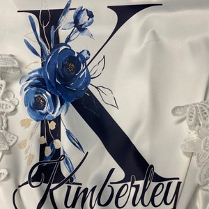 May include: White satin robe with a navy blue letter K adorned with blue and gold floral designs. The name Kimberly is written in cursive below the letter.