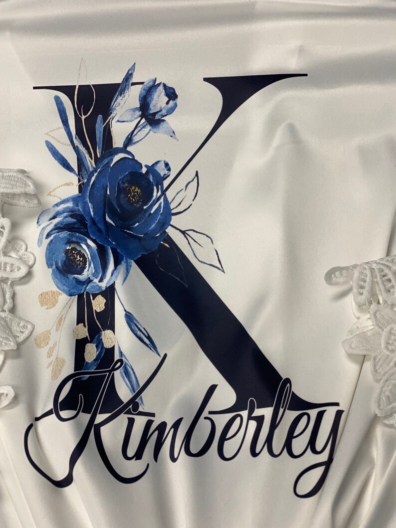 May include: White satin robe with a navy blue letter K adorned with blue and gold floral designs. The name Kimberly is written in cursive below the letter.