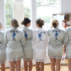 May include: Five women wearing matching satin robes with lace trim. The robes are personalized with a large initial and a floral design. The robes are in shades of white and light blue.