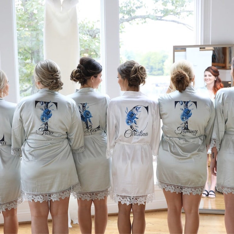 May include: Five women wearing matching satin robes with lace trim. The robes are personalized with a large initial and a floral design. The robes are in shades of white and light blue.