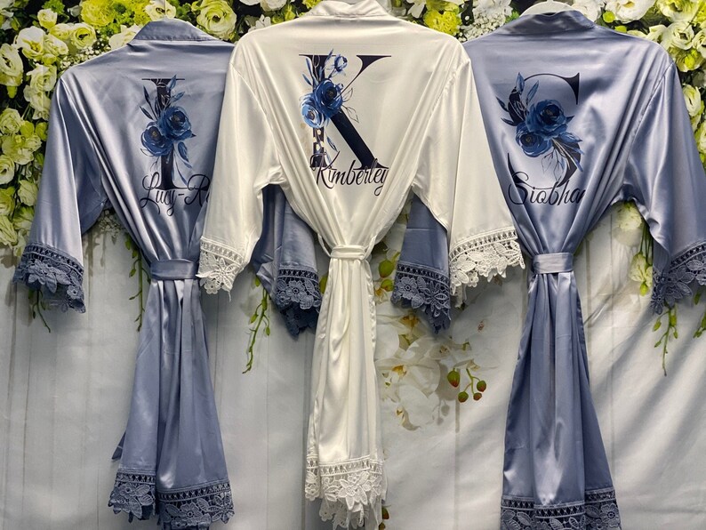 May include: Three satin robes with lace trim. The robes are in shades of blue and white. The robes are personalized with a letter and a name. The names are Lucy-R, Kimberly, and Siobha.