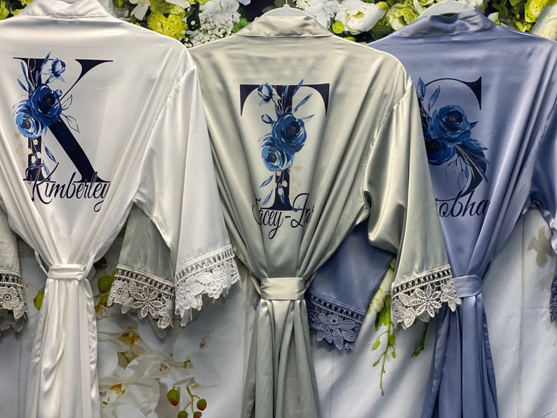 May include: Three white, gray, and blue satin robes with lace trim and personalized with a floral initial and name. The names are Kimberly, Lacey-Jo, and Sophia.