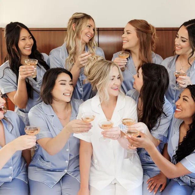 May include: A group of women wearing matching light blue pajamas raise their champagne glasses in a toast. They are smiling and looking at each other. One woman is wearing a white robe.