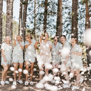 May include: A group of women in white pajamas celebrate with a champagne toast in a forest setting. The women are smiling and laughing as they pop the champagne bottle, creating a shower of bubbles.