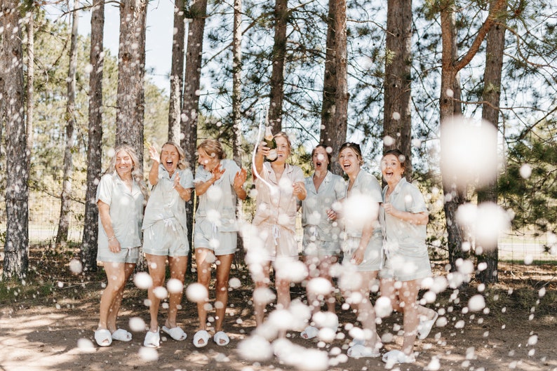May include: A group of women in white pajamas celebrate with a champagne toast in a forest setting. The women are smiling and laughing as they pop the champagne bottle, creating a shower of bubbles.