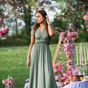 Peut inclure: Une femme portant une robe maxi vert sauge &agrave; col en V et manches courtes se tient dans un jardin. La robe est de style portefeuille et est cintr&eacute;e &agrave; la taille. La femme est entour&eacute;e de fleurs roses et de verdure.