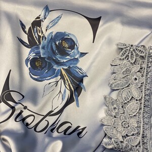 May include: A light blue satin robe with a floral design and the name "Siobhan" printed on the front. The robe has lace trim on the sleeves.