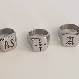 May include: Three silver signet rings with different engravings. The first ring has the initials "A.S." engraved on it. The second ring has the initials "S.T." engraved on it, with three small hearts between the letters. The third ring has the letter "A" engraved on it in a gothic font.