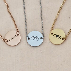 Puede incluir: Tres collares de plata con colgantes redondos. Los colgantes tienen diferentes grabados: uno dice "12.07.14", otro tiene un diseño de dos manos haciendo una promesa meñique, y el tercero dice "E & D" con un símbolo de corazón entre las letras.