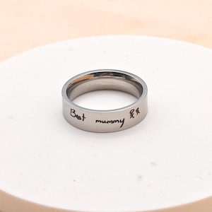 Puede incluir: Un anillo plateado con las palabras "Best mummy" y una peque&ntilde;a ilustraci&oacute;n de dos figuras. El anillo est&aacute; sobre un fondo claro.