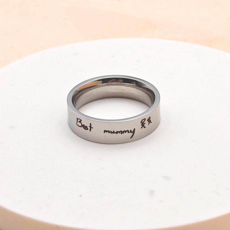 Puede incluir: Un anillo plateado con las palabras "Best mummy" y una peque&ntilde;a ilustraci&oacute;n de dos figuras. El anillo est&aacute; sobre un fondo claro.