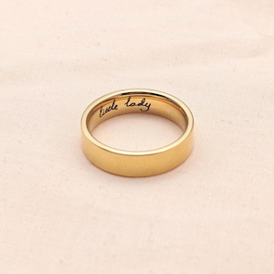 Puede incluir: Un anillo dorado con la inscripci&oacute;n "little lady" grabada en el interior. El anillo tiene una superficie lisa y pulida y un dise&ntilde;o de banda cl&aacute;sico. El anillo est&aacute; sobre un fondo rosado suave.
