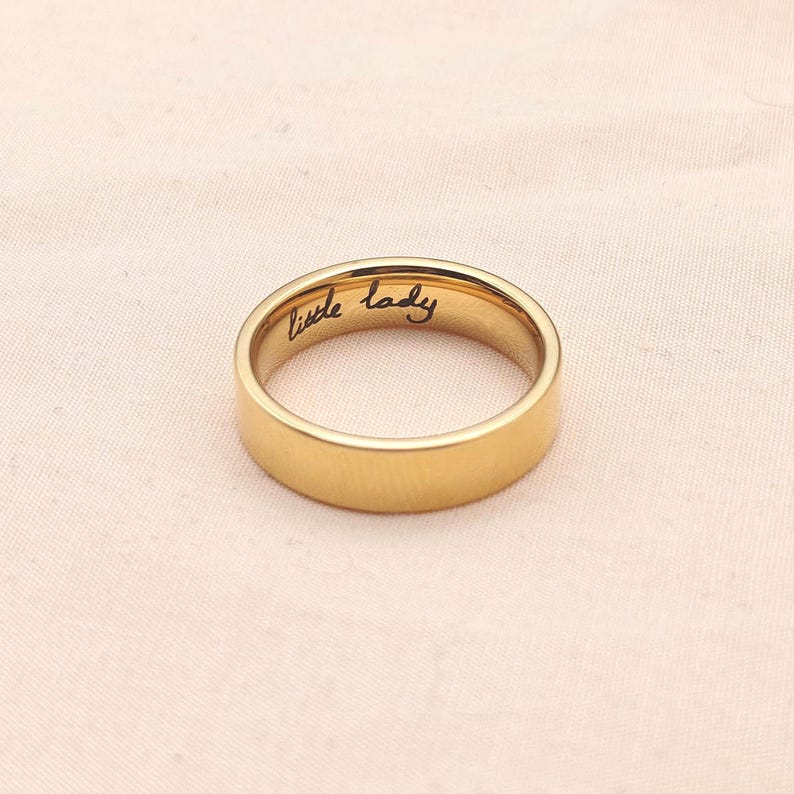 Puede incluir: Un anillo dorado con la inscripci&oacute;n "little lady" grabada en el interior. El anillo tiene una superficie lisa y pulida y un dise&ntilde;o de banda cl&aacute;sico. El anillo est&aacute; sobre un fondo rosado suave.