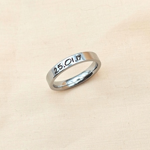Puede incluir: Un anillo plateado con la fecha "25.01.19" grabada. El anillo es liso y reflectante, sobre una tela rosa claro. El anillo es de dise&ntilde;o sencillo, con forma de banda.