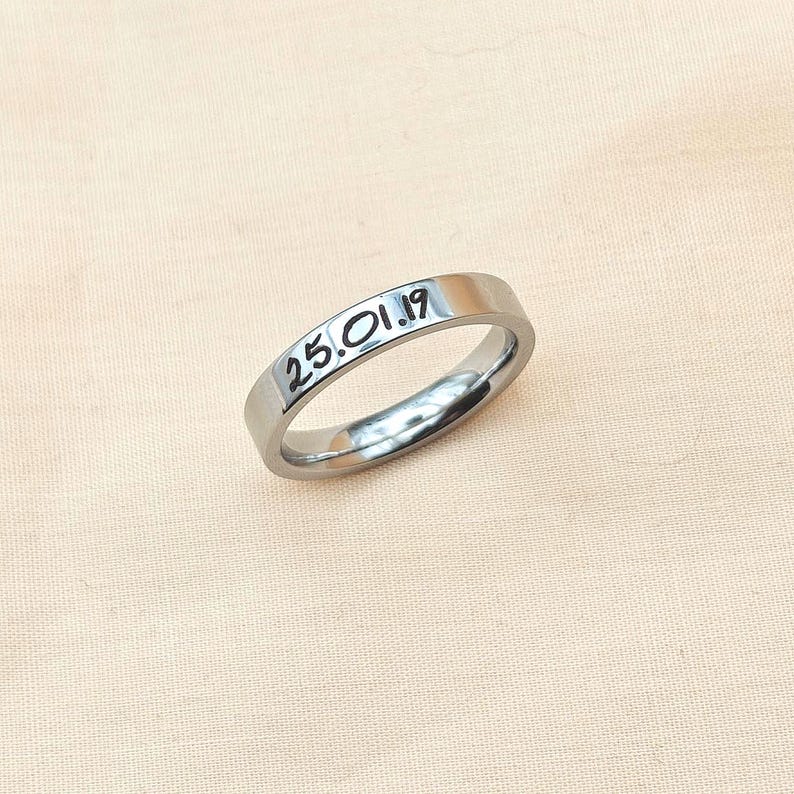 Puede incluir: Un anillo plateado con la fecha "25.01.19" grabada. El anillo es liso y reflectante, sobre una tela rosa claro. El anillo es de dise&ntilde;o sencillo, con forma de banda.