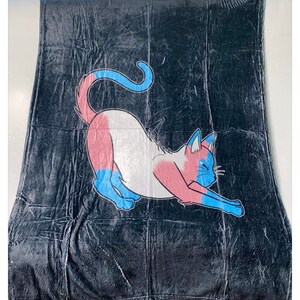 May include: A black fleece blanket with a cartoon cat design. The cat is white with pink, blue, and light blue accents.