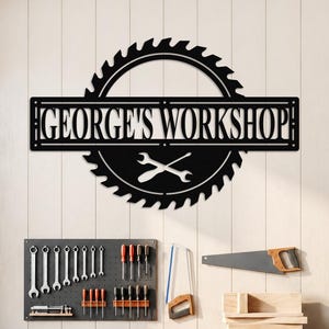 Op de afbeelding: Zwart metalen bord met de tekst "GEORGE'S WORKSHOP" boven een zaagblad en gekruiste sleutels. Het bord is gemonteerd op een lichtgekleurde muur boven een houten werkbank met gereedschap. Een pegboard met gereedschap is aan de muur bevestigd.