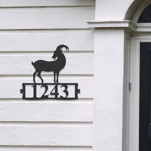 Puede incluir: Placa de número de casa de metal negro con una silueta de cabra de montaña y el número 1243. La placa está montada en una pared blanca junto a una puerta negra.