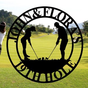 Puede incluir: Un letrero circular de metal negro con el texto "John & Floras 19th Hole" en una fuente decorativa. El letrero presenta siluetas de dos golfistas, un hombre y una mujer, sosteniendo palos de golf.