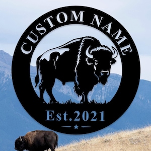 May include: Black metal bison wall art with the words "CUSTOM NAME" in a circular design. The text "Est. 2021" is below the bison. The background shows a mountain range and a bison on a grassy hill.