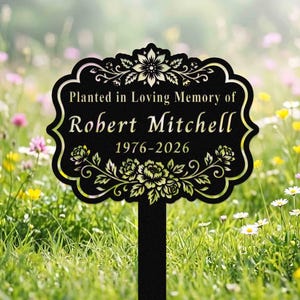 May include: A black metal garden memorial stake with a decorative scalloped edge, featuring the engraved text "Planted in Loving Memory of Robert Mitchell 1976-2026" and ornate floral motifs at the top and bottom of the rectangular plaque.