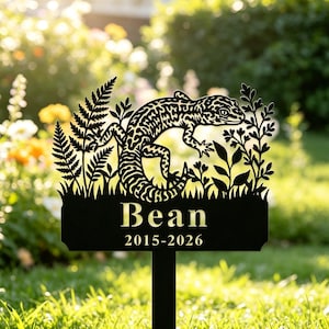Puede incluir: Un estaca de jardín de metal negro con una silueta recortada de un gecko leopardo rodeado de follaje. La base rectangular muestra el nombre Bean en letras doradas sobre los años 2015-2026, también en dorado, sobre un fondo negro.