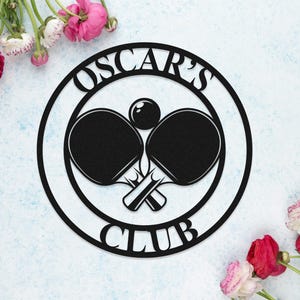 Puede incluir: Un letrero de metal negro con el texto "Oscar's Club" en un círculo. Dentro del círculo hay dos palas de ping pong cruzadas y una pelota.