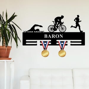 Könnte beinhalten: Schwarzer Medaillenhalter aus Metall mit drei Reihen Haken. Der Halter zeigt eine Silhouette eines Schwimmers, Radfahrers und Läufers. Der Name "BARON" ist auf dem Halter aufgedruckt. Zwei Goldmedaillen hängen an den Haken.