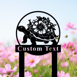 Puede incluir: Estaca de jardín de metal negro con una silueta redonda de una tortuga con flores en su espalda. La estaca tiene un letrero rectangular que dice "Texto personalizado".