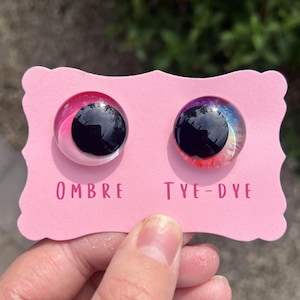 Pode incluir: Dois cabochões redondos, um com efeito degradê rosa e outro com padrão tie-dye, são exibidos em um cartão rosa. As palavras "OMBRE" e "TYE-DYE" são impressas abaixo. Os cabochões são segurados por uma mão.