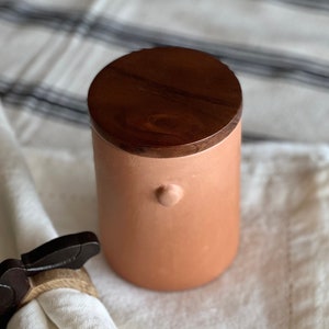 May include: A white linen napkin with a wooden napkin ring and a terracotta jar with a wooden lid.