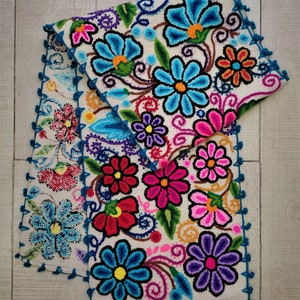 May include: Two colorful embroidered table runners with a floral design. The runners are white with blue, pink, red, yellow, and purple flowers. The edges of the runners are trimmed with blue crocheted edging.