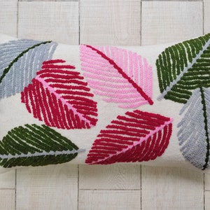 Peut inclure: Un coussin blanc avec un motif brodé coloré de feuilles dans des tons de rose, rouge, vert et gris.