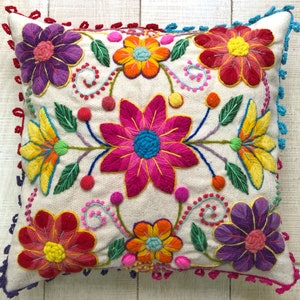 May include: A white pillow with a colorful embroidered floral design. The pillow has a fringe border and features pink, yellow, orange, purple, and green flowers.