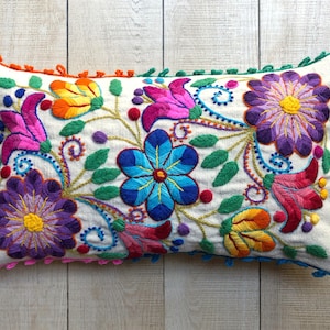May include: A white pillow with a colorful floral embroidery design. The pillow has a fringed border in orange, green, and pink.