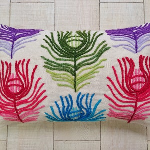 Peut inclure: Un coussin rectangulaire blanc avec des plumes de paon brodées colorées. Les plumes sont dans des tons de violet, vert, rose et bleu.
