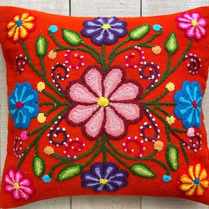 Può includere: Un cuscino quadrato rosso con disegni floreali ricamati in rosa, blu, giallo e viola. Il cuscino ha uno stile messicano tradizionale.