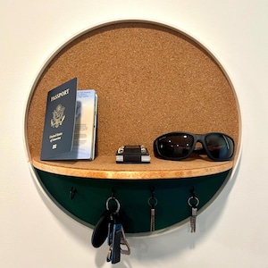 Puede incluir: Un organizador de pared redondo con respaldo de corcho, un estante de madera y una sección inferior verde oscuro. Contiene un pasaporte, gafas de sol, una cartera pequeña y llaves. El organizador tiene un marco blanco y ganchos para llaves.