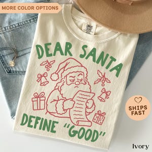 May include: An ivory t-shirt featuring a red line art graphic of Santa Claus reading a list, surrounded by red bells, gifts, and holly. The text in green reads "DEAR SANTA DEFINE "GOOD"". The shirt has a Comfort Colors brand label at the neck.