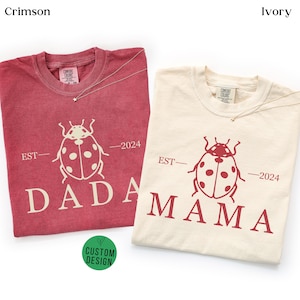 May include: Two t-shirts, one crimson and one ivory, each with a ladybug graphic and the words "DADA" and "MAMA" printed in a bold font. The shirts also feature the text "EST-2024". A small green circle with the words "CUSTOM DESIGN" is visible.