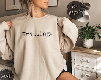 May include: A sand-colored sweatshirt with the word "Knitting." printed in black. The long-sleeved crewneck top is made of a soft material. The sweatshirt is part of a clothing collection.