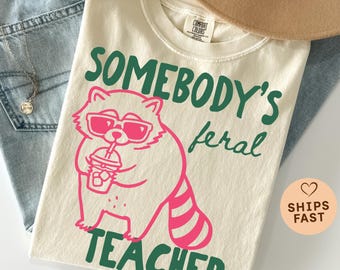 May include: Ivory-colored t-shirt with a raccoon graphic wearing sunglasses and holding a drink. The text reads "SOMEBODY'S feral TEACHER" in green and pink. The shirt is folded on top of blue jeans.