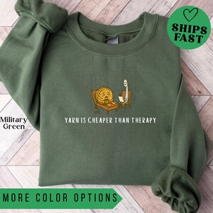 May include: Military green sweatshirt with the text "YARN IS CHEAPER THAN THERAPY" and a cartoon of a ball of yarn in a chair with a therapist. The sweatshirt also has the words "MORE COLOR OPTIONS" and a green heart that says "SHIPS FAST".