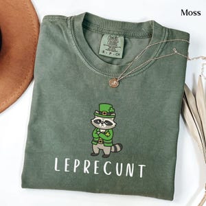 May include: Moss green t-shirt with a graphic of a raccoon dressed as a leprechaun, with the word "LEPRECUNT" printed below. The shirt has a crew neck and short sleeves. A gold necklace with a pendant is draped on the shirt.